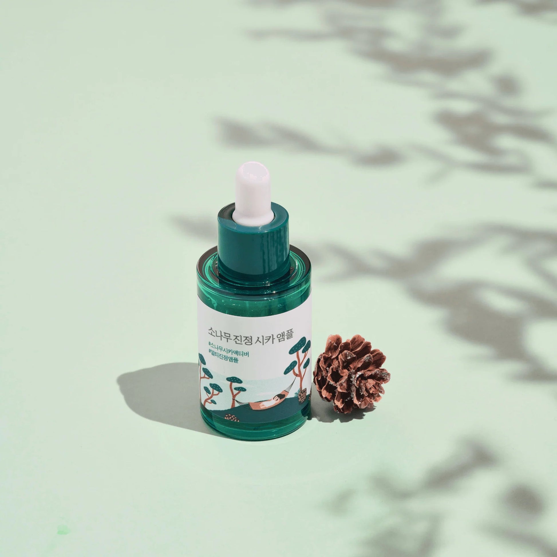 Round Labs Pine Tree Calming Cica Ampoule on a green background with pine cone next to it