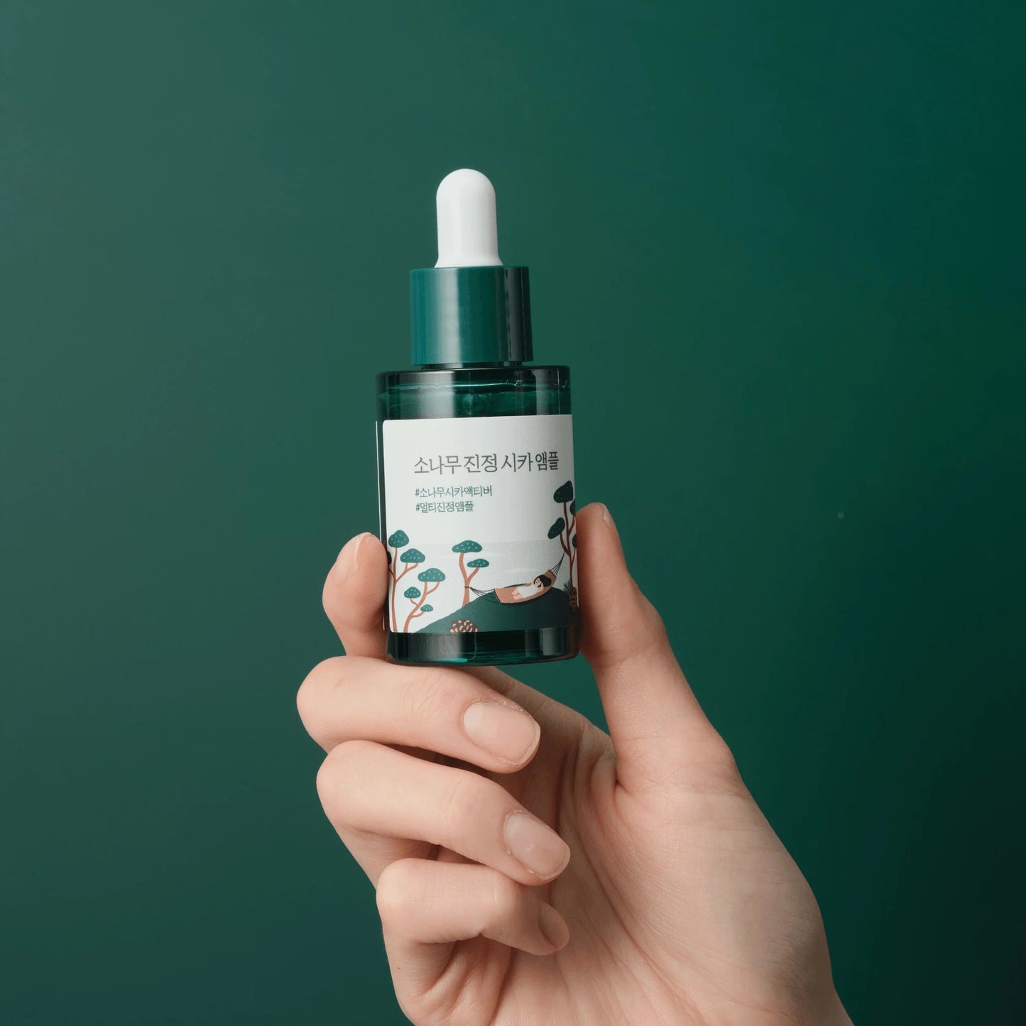 Round Labs Pine Tree Calming Cica Ampoule being held by a hand on dark green background